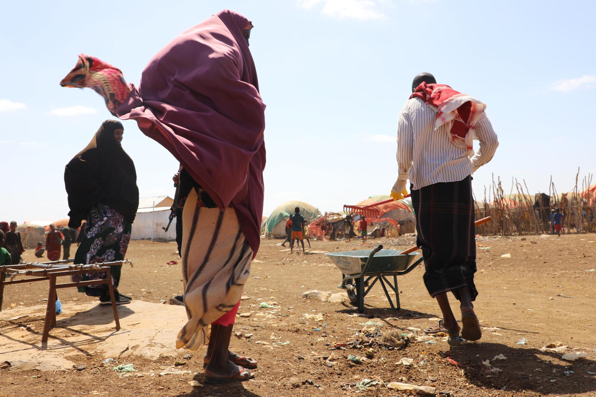 A man with a rake and wheelbarrow and women in wind-blown shawls at a settlement of makeshift shelters, with children playing in the background